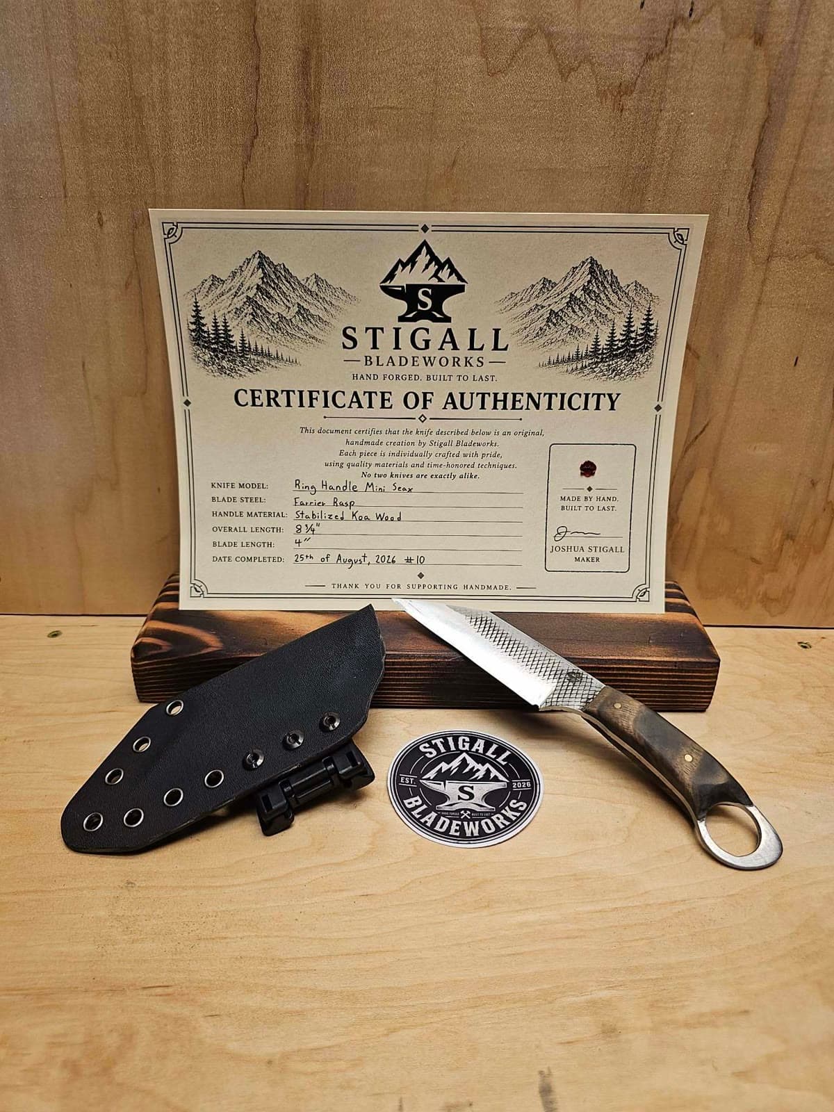 Ring-handle mini seax with a rasp-textured blade and koa handle beside its Kydex sheath and a Stigall Bladeworks sticker, in front of its signed certificate of authenticity